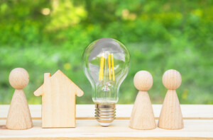 Wooden house, lightbulb and many little men on a wooden table. Energy saving, debts for utility bills.
