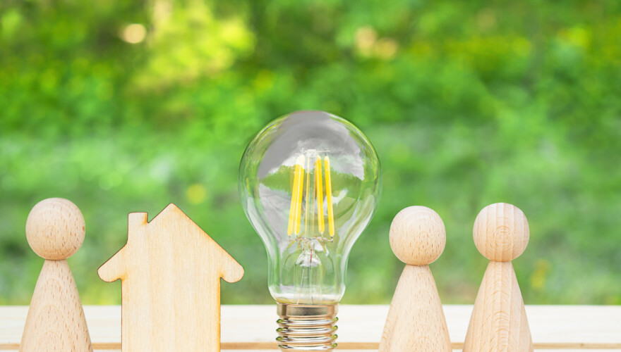 Wooden house, lightbulb and many little men on a wooden table. Energy saving, debts for utility bills.