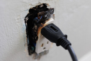A close-up of a burnt electrical outlet with a plug still inserted, showing signs of damage from an electrical fire caused by faulty wiring.