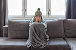 Cold Home. Young Woman Covered With Blanket Freezing On Couch In Living Room, Wearing Knitted Scarf And Hat Indoors Warming Hands With Her Breath, Suffering Heating Problems