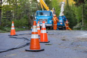 Selective focus view of high viz road cones warning traffic of fallen overhead power line after storm. In the background, blurry contractors work to repair power lines and restore electricity after a storm.