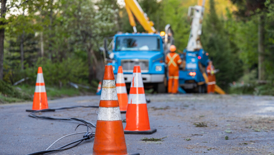 Selective focus view of high viz road cones warning traffic of fallen overhead power line after storm. In the background, blurry contractors work to repair power lines and restore electricity after a storm.