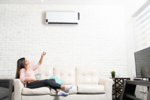 Latin woman turning on air conditioner unit using remote control while sitting on sofa at home