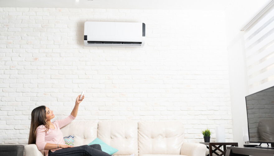 Latin woman turning on air conditioner unit using remote control while sitting on sofa at home