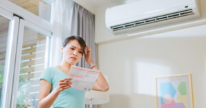 Asian woman stand by air conditioner shocked and upset about electricity bill in hand at home