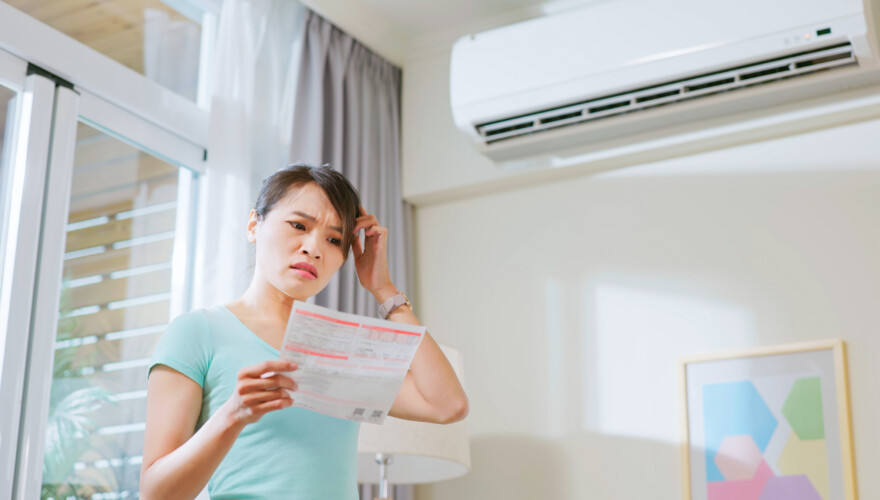 Asian woman stand by air conditioner shocked and upset about electricity bill in hand at home