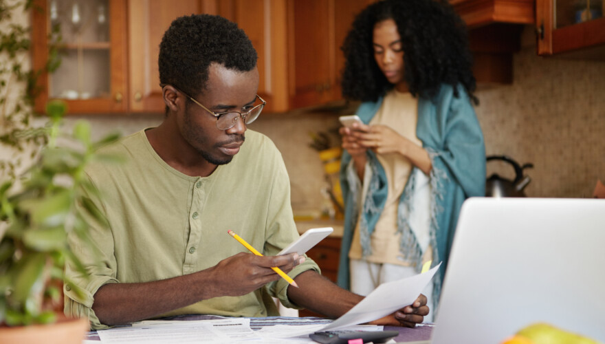 A dark-skinned male holding a mobile phone in one hand and piece of paper in other, comparing energy services