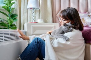 Woman sitting on floor with cat under warm blanket warming near an electric heater. Middle-aged female with pet at home in bedroom, heating up near heating radiator.