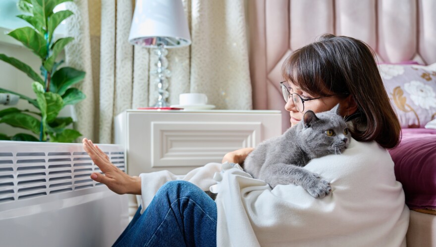 Woman sitting on floor with cat under warm blanket warming near an electric heater. Middle-aged female with pet at home in bedroom, heating up near heating radiator.
