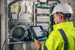 An electrical technician using a tablet while working in a switchboard with fuses.
