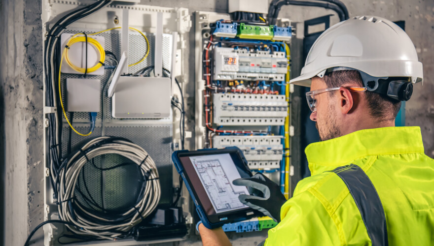 An electrical technician using a tablet while working in a switchboard with fuses.